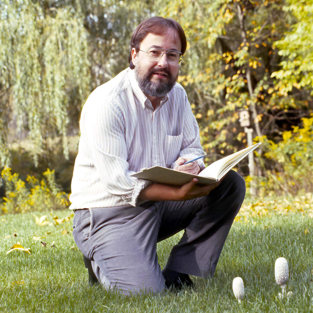 Glen Loates outside by mushrooms kneeling with sketchbook and smiling